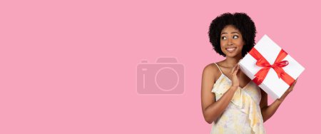Photo for Cheerful african american young woman with curly hair, pleasantly surprised, holding a large white gift box with a vibrant red ribbon, on a soft pink background, expressing happiness and excitement - Royalty Free Image