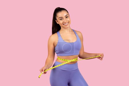Photo for Cheerful fit woman in lilac gym outfit proudly displaying measuring tape around her waist, symbolizing weight loss success on pink background - Royalty Free Image