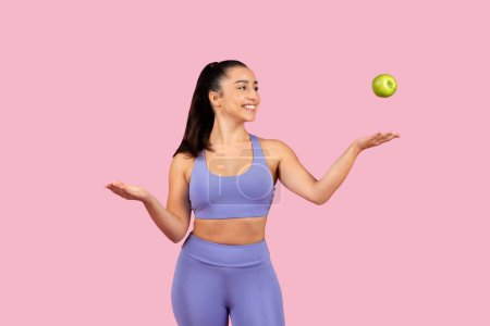 Photo for Smiling, active woman in athletic wear playfully tossing green apple in the air against vibrant pink background, representing healthy lifestyle and sport - Royalty Free Image