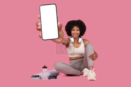 Téléchargez les photos : Femme noire souriante en tenue d'entraînement s'assoit jambes croisées, affichant un écran de smartphone vide alors qu'elle est entourée de vêtements de fitness, écouteurs autour de son cou, sur fond rose, maquettes - en image libre de droit