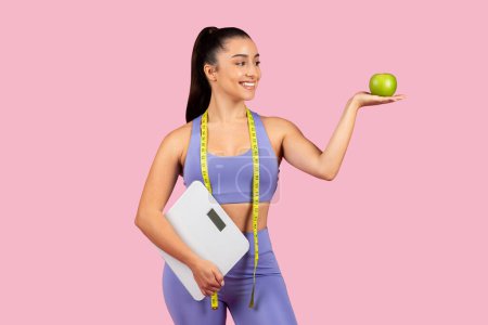 Téléchargez les photos : Femme axée sur la condition physique tenant la balance et la pomme verte sur sa paume, posant avec du ruban à mesurer autour de son cou, représentant l'alimentation et la santé - en image libre de droit