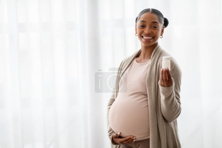 Photo for Cheerful young african american expecting woman hugging her big belly and holding white jar with pills, standing next to window at home. Multivitamins, supplements during pregnancy, copy space - Royalty Free Image