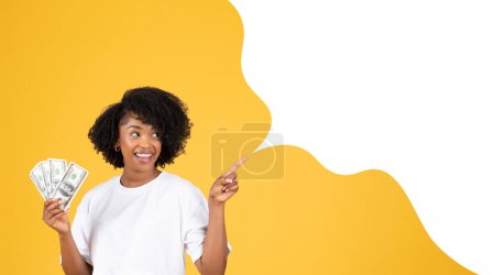 Foto de Oferta financiera. Mujer afroamericana feliz sosteniendo dinero en efectivo en dólares y señalando con el dedo la burbuja del habla vacía con burla para publicidad, sobre fondo amarillo. Collage, Panorama - Imagen libre de derechos