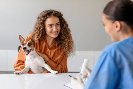 Téléchargez les photos : Femme aux cheveux bouclés sourit tout en tenant son Jack Russell Terrier ludique comme ils consultent un vétérinaire féminin sur la santé des chiens à la clinique - en image libre de droit