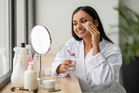 Téléchargez les photos : Jeune femme indienne positive portant un peignoir blanc nettoyant visage dans la salle de bain, en utilisant du coton et de l'eau micellaire, démaquillant, regardant miroir et souriant - en image libre de droit