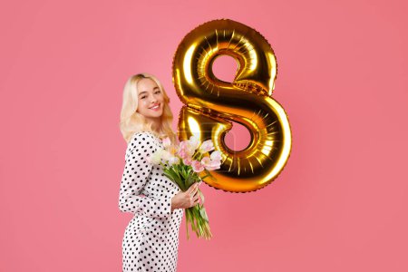 Photo for Smiling blonde woman in dotted dress holds pink tulips and golden number 8 balloon, celebrating Womens International Day on pink background - Royalty Free Image