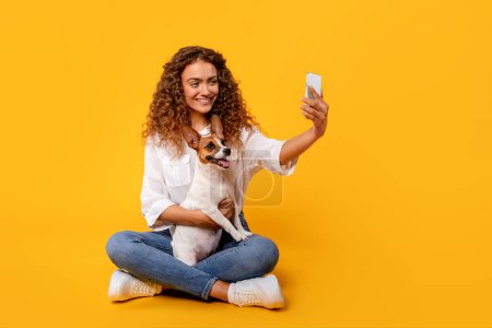 Téléchargez les photos : Femme aux cheveux bouclés capture joyeusement moment selfie avec son excité Jack Russell Terrier, assis sur fond jaune vibrant, espace libre - en image libre de droit