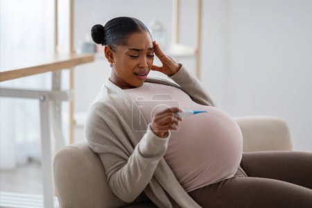 Fieber während der Schwangerschaft. Schwangere Afroamerikanerin mit Thermometer zur Temperaturmessung fühlt sich zu Hause auf Sofa schlecht an Leerer Raum, Seitenblick