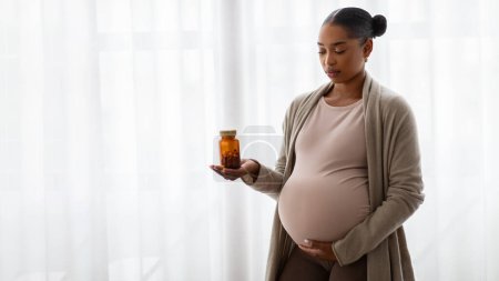 Photo for Pregnant young african american lady hugging her big belly and holding brown jar with pills, standing next to window at home. Multivitamins, supplements during pregnancy, panorama, copy space - Royalty Free Image