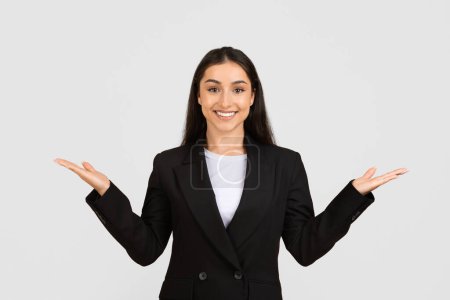 Photo for Smiling professional woman in business suit stands with her hands open in welcoming, presenting gesture, posing on light grey background - Royalty Free Image