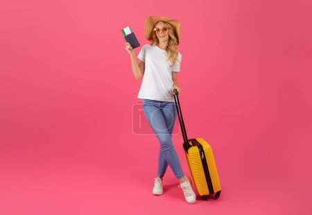 Photo for Ready for Summer Getaway. Blonde woman in straw hat with sunglasses and luggage, holding passport and boarding pass, ready to travel while standing on pink background. Full length, copy space - Royalty Free Image