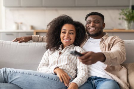 Téléchargez les photos : Temps libre à la télé. Couple afro-américain souriant regardant la télévision ensemble à l'intérieur de la maison, mari tenant et pointant télécommande commutant les chaînes de télévision. Confort domestique et plaisir - en image libre de droit