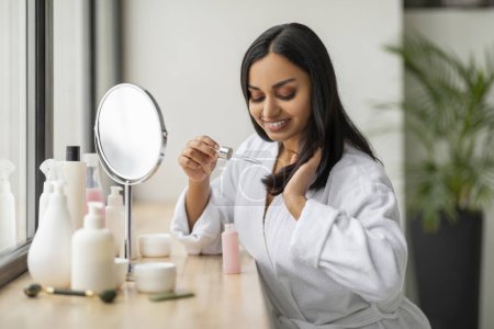 Foto de Concepto de cuidado del cabello. Hermosa mujer india milenaria con albornoz blanco sentado frente al espejo en el baño, usando aceite para el cabello termina el tratamiento, disfrutando de la rutina de la mañana - Imagen libre de derechos