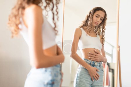 Téléchargez les photos : Adolescente malheureuse touchant son estomac devant un miroir à l'intérieur de la maison. Concept représentant des maux d'estomac pendant les crampes menstruelles, problèmes de santé gynécologique, mécontentement sur la forme du corps - en image libre de droit