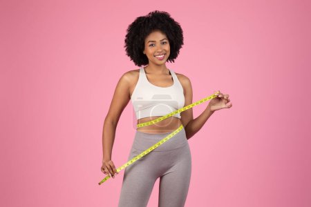 Téléchargez les photos : Souriante jeune femme noire mesure avec confiance sa taille avec du ruban à mesurer jaune, démontrant une image corporelle saine et des progrès de forme physique, sur fond rose - en image libre de droit