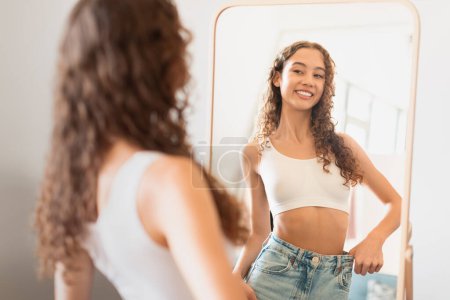 Photo for Happy teen girl at home looks at mirror reflection satisfied with weight loss indoor, comparing slim waist wearing oversized jeans after diet and slimming, embracing healthier lifestyle - Royalty Free Image