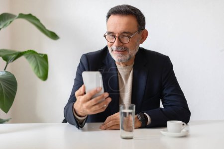 Foto de Hombre de negocios caucásico mayor en una chaqueta y vasos mira a su teléfono inteligente con interés, sentado en una mesa blanca con una taza de café y un vaso de agua. Trabajo, aplicación de negocios, videollamada - Imagen libre de derechos