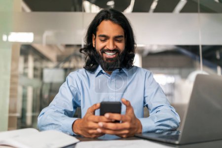 Photo for Business application concept. Positive indian guy in formal outwear manager using smartphone, sitting at desk in front of laptop computer, reading email, modern office interior - Royalty Free Image