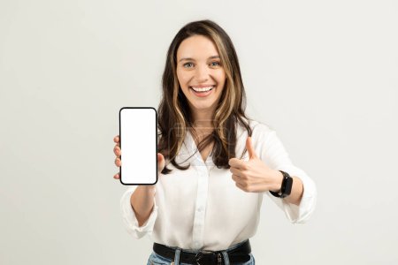 Téléchargez les photos : Jeune femme européenne enthousiaste avec un sourire éclatant, montrant un écran de smartphone vierge d'une main et donnant un pouce vers le haut avec l'autre, sur un fond clair, studio. Travail, entreprise - en image libre de droit