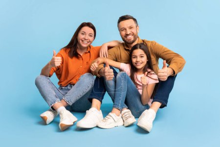 Photo for Family Approval. Cheerful parents and kid daughter making thumbs up gesture together, sitting on floor on blue studio background, smiling to camera approving offer, full length. - Royalty Free Image