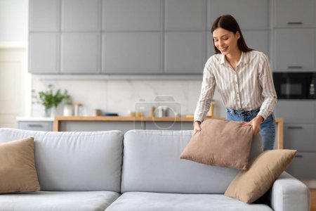 Foto de Mujer joven con expresión alegre organiza cuidadosamente cojines suaves en elegante sofá gris en un espacio de vida bien iluminado y contemporáneo, lo que indica sentido de orgullo en su acogedora casa - Imagen libre de derechos