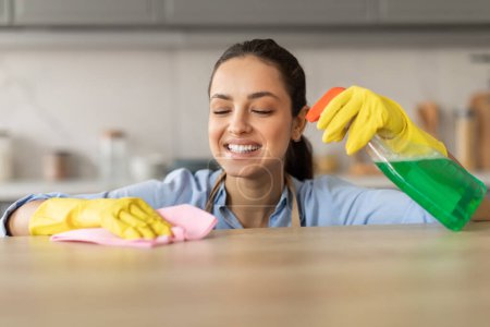 Foto de Mujer en camisa azul y guantes amarillos alegremente usando aerosol botella y tela para limpiar la encimera de la cocina, disfrutando de las tareas domésticas, primer plano - Imagen libre de derechos