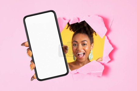 Téléchargez les photos : Heureuse jeune femme afro-américaine excitée avec la bouche ouverte montrant téléphone portable avec écran vide à travers le trou dans le papier, fond de studio rose. Site web, app, recommandation, soins de beauté - en image libre de droit