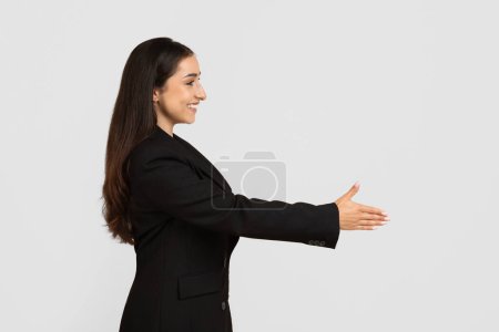 Photo for Confident businesswoman with long hair in formal black suit extending her hand for friendly handshake, showcasing professional greeting, side view, free space - Royalty Free Image