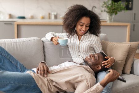 Foto de Acogedor ambiente de fin de semana. Relajada pareja afroamericana comparte momentos tiernos en la sala de estar, abrazándose en el sofá con una taza de té y un mando a distancia, creando un fondo de amor y felicidad - Imagen libre de derechos