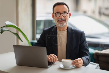 Foto de Hombre mayor caucásico accesible con una sonrisa agradable trabajando en un ordenador portátil en un café, disfrutando de una taza de café, creando una atmósfera de facilidad productiva. Trabajo, negocios - Imagen libre de derechos
