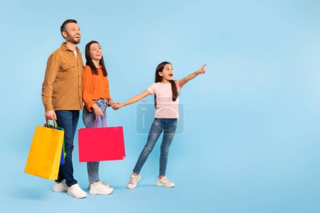 Photo for Joyful family after shopping with daughter holding paper shopper bags, kid girl pointing finger at empty space for offer, advertising nice commercial deal over blue backdrop, full length - Royalty Free Image