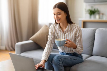 Photo for Cheerful young woman enjoying cup of coffee while using her laptop, comfortably sitting on sofa in cozy and well-lit living room interior, lady browsing internet or working remotely - Royalty Free Image
