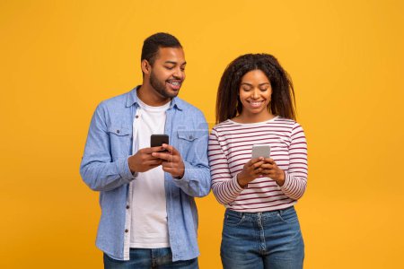 Photo for Cheerful young black couple using their smartphones, happy african american man and woman sharing moment of digital connection with warm smiles, standing against yellow studio background - Royalty Free Image