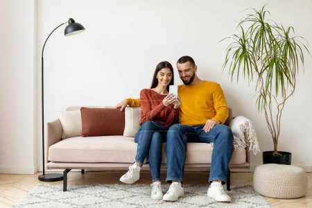 Photo for Young Smiling Caucasian Couple Using Mobile Phone And Embracing Together At Home, Happy Millennial Spouses Relaxing On Couch With Modern Smartphone, Browsing New App Or Shopping Online, Copy Space - Royalty Free Image