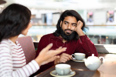 Téléchargez les photos : Un rencard raté. Jeune homme indien barbu se sentant ennuyé au déjeuner au café, regardant sa petite amie partager des pensées avec l'expression du visage déçu, foyer sélectif - en image libre de droit
