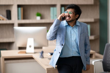 Photo for Smiling middle eastern businessman discussing details over phone looking away at modern office interior. Modern business communication concept. Free space for text and mobile offers - Royalty Free Image