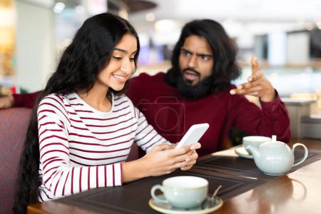 Foto de Hombre indio joven enojado haciendo gestos y gritando, tener pelea con la novia sonriendo en la pantalla del teléfono inteligente mientras cita romántica en la cafetería. Pareja oriental que experimenta dificultades en las relaciones - Imagen libre de derechos