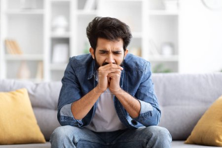 Photo for Sorrow And Grief. Depressed young indian man sitting on sofa in living room at home, upset eastern male looking down with deeply troubled expression, expressing negative emotions, copy space - Royalty Free Image