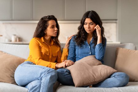 Photo for Attentive young woman comforting depressed friend or sister holding her hand, providing support to upset girlfriend in difficult life situation, helping cope with problems, sitting on couch indoor - Royalty Free Image