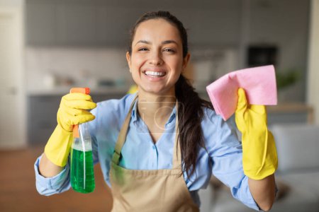 Téléchargez les photos : Femme aux yeux brillants en gants jaunes tenant le flacon de pulvérisation vert et la lingette rose, prête et heureuse de s'attaquer aux tâches ménagères - en image libre de droit