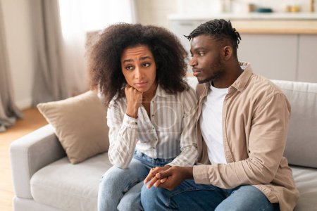 Relationship struggle. Unhappy African American couple holding hands, navigating issues of indifference and betrayal, sitting on couch together in modern living room. Love challenges