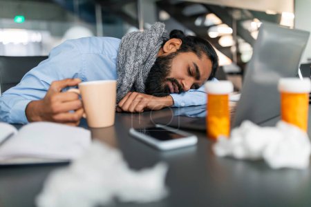Téléchargez les photos : Homme d'affaires surmené homme hindou millénaire dans une chemise bleue et écharpe endormi à son bureau, entouré de pots avec des pilules et des tissus, tenant tasse avec boisson chaude, gars malade travaillant au bureau - en image libre de droit