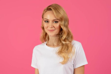 Téléchargez les photos : Portrait de jolie jeune femme blonde en t-shirt blanc regardant avec un sourire mignon à la caméra, posant avec des cheveux lâches sur fond de studio rose, la publicité beauté ou l'offre de mode - en image libre de droit