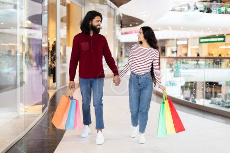 Photo for Positive young eastern spouses have family shopping at mall. Cheerful indian man and woman holding hands and walking by store, carrying colorful paper bags purchases. Season sale - Royalty Free Image