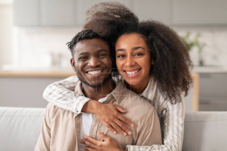 Téléchargez les photos : Portrait d'un couple afro-américain souriant partageant une tendre étreinte sur un canapé à la maison, regardant la caméra. La femme aimante embrasse son mari. Le bonheur et l'amour entre deux conjoints concept - en image libre de droit