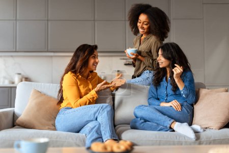 Foto de Estudiantes multiétnicas mujeres comparten historias y sonrisas, sentarse con tazas de té en fin de semana tranquilo, hablar y divertirse en la cómoda sala de estar moderna. Novia trío charlando sobre café - Imagen libre de derechos