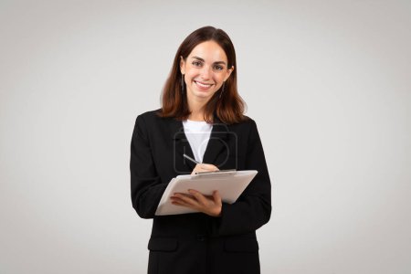 Téléchargez les photos : Femme d'affaires européenne souriante et confiante, au sourire chaleureux, écrivant sur une tablette numérique, vêtue d'un costume formel, incarnant professionnalisme et efficacité, studio - en image libre de droit