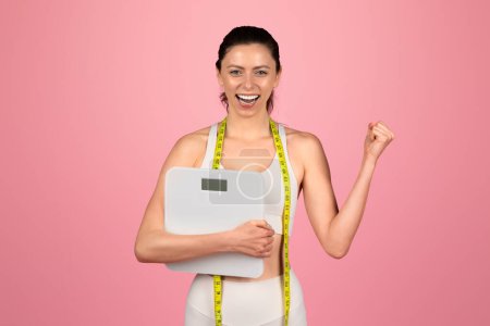 Photo for Exultant european young woman celebrating her weight loss success, holding a scale and wearing a measuring tape as a sash, feeling triumphant on a pink background, studio - Royalty Free Image
