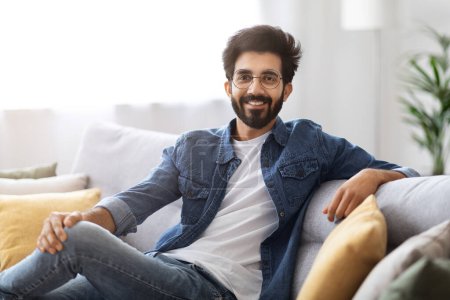 Photo for Portrait Of Smiling Young Indian Man Posing On Couch At Home, Handsome Millennial Eastern Guy In Casual Clothes Resting On Sofa In Living Room And Looking At Camera, Enjoying Domestic Leisure - Royalty Free Image