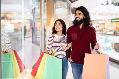 Photo for Family Shopping. Excited Indian Couple Holding Colorful Shopper Bags, Embracing Posing In Modern Mall Indoors. Seasonal Sales, Great Offer Concept. Selective Focus On Spouses - Royalty Free Image